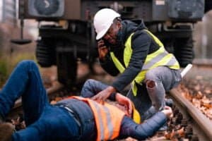 imagem mostra uma cena de acidente, com um trabalhador no chão, outro trabalhador ligando para emergência e ao fundo trilhos e um trem.